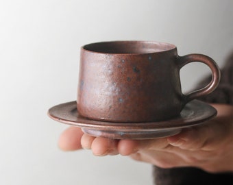 May include: A brown ceramic coffee cup and saucer with a speckled glaze. The cup has a rounded shape and a handle. The saucer is round and has a slightly raised edge.
