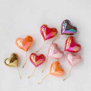 May include: A collection of nine iridescent heart-shaped balloons in various colours, including gold, pink, red, and orange. The balloons are tied with gold string and are arranged on a white surface.