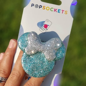 May include: A glittery PopSockets phone grip in the shape of a bow, with a silver bow and turquoise ears. The PopSockets packaging is visible in the background, with the brand name and a phone graphic.