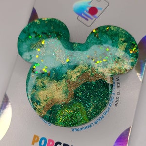 May include: A teal and gold Mickey Mouse-shaped phone grip. The grip is made of resin and contains gold flakes and glitter. The grip is on a white backing with the PopGrip logo.