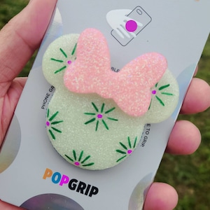 May include: A PopSockets PopGrip featuring a Minnie Mouse design. The grip has a light green base with green and pink floral accents, and a pink glitter bow. The packaging is white with the PopSockets logo.