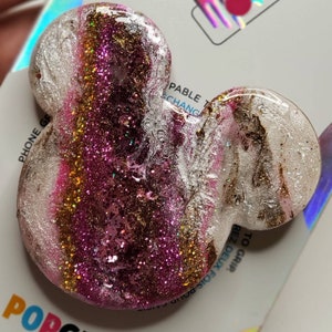 May include: A glittery, resin PopGrip in the shape of Mickey Mouse ears. The design features layers of pink, gold, and silver glitter. The PopGrip is attached to a white backing card with the brand name "PopGrip" visible.