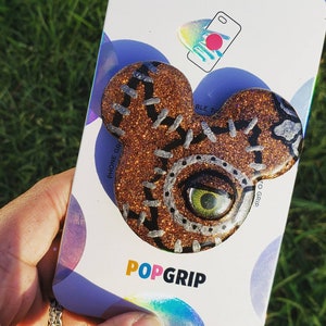 May include: A glittery brown PopSocket phone grip in the shape of a mouse head with a stitched design and a green eye. The PopSocket is attached to a white backing with the PopSockets and PopGrip logos.