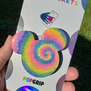 May include: A PopSockets PopGrip in the shape of Mickey Mouse ears, featuring a colorful tie-dye swirl design with glitter accents. The PopGrip is attached to a white and silver PopSockets branded backing. The words "POPSOCKETS" and "POPGRIP" are printed on the backing.