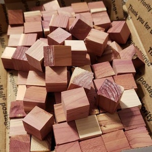 May include: A cardboard box filled with numerous small, square cedar wood blocks. The blocks vary in color from light tan to deep reddish-brown, showcasing the natural wood grain. The box is open, revealing the contents.