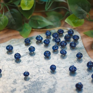 May include: A collection of small, dark blue ceramic blueberries arranged on a silver metal surface. The blueberries are scattered across the surface, creating a visually appealing display.