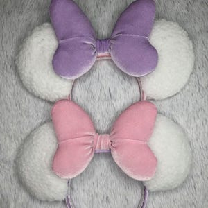 May include: Two headbands with plush white ears and velvet bows. One bow is lavender, the other is pink. The headbands have a thin, decorative band. The background is a soft, textured gray.