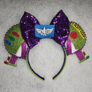 May include: A Buzz Lightyear-themed headband with a purple sequined bow and green and purple accents. The headband features Buzz Lightyear's wings and the words "LIGHTYEAR". The design includes a blue emblem with a white wing design.