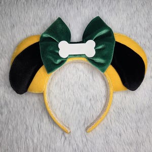 Può includere: Una fascia per capelli a tema cane con orecchie gialle, dettagli neri sulle orecchie e un fiocco di velluto verde. Un accento bianco a forma di osso è centrato sul fiocco. La fascia è gialla.