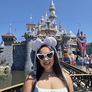 May include: A person wearing heart-shaped sunglasses and a white bow headband with a veil, posing in front of a large castle with blue and gold spires. The person is wearing a white dress and a necklace.