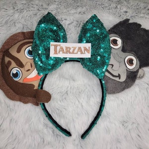 May include: Green sequin Minnie Mouse ears with a brown and gray felt character head on each side. The character on the left is Tarzan and the character on the right is Terk. The bow has a gold glitter tag that says "TARZAN".