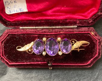 May include: A gold brooch with three oval amethyst stones and pearl accents. The brooch is in a red velvet box with a label that says "Box N/F Sale".