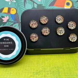 May include: A set of magnetic pins with animal print designs, including snakeskin and leopard patterns. The pins are displayed on a black metal container and a black magnetic surface. A round tin with the text "PIN DREAMS ON - MAGNETIC PINS" is also in the image.
