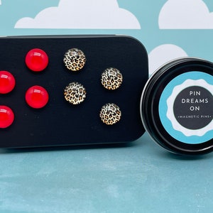 May include: A set of six magnetic pins in a black tin. The pins are red and have a leopard print design. The tin has the text "Pin Dreams On - Magnetic Pins" on a blue and white label.