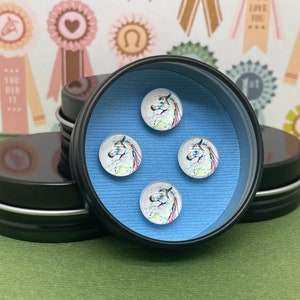 Könnte beinhalten: Vier runde Glascabochons mit bunten, bemalten Pferde-Designs werden in einer schwarzen Dose mit blauem Hintergrund präsentiert.
