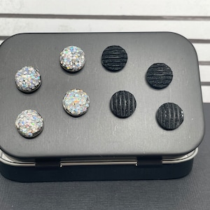 May include: A black tin box with six round buttons. Three buttons are black with a textured surface and three buttons are iridescent with a sparkly finish.