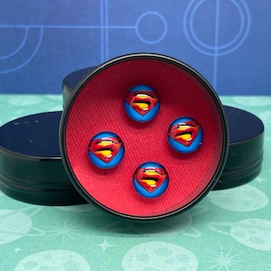 May include: Four round glass buttons with a Superman logo on a red background inside a black round container.