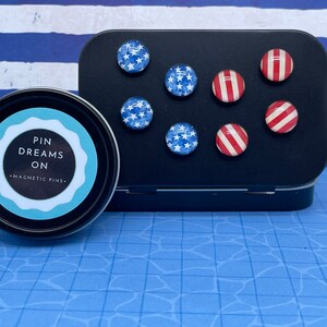 May include: A set of magnetic pins with patriotic designs, including blue stars on a white background and red and white stripes. The pins are displayed on a black metal container and in a round tin with the text "PIN DREAMS ON - MAGNETIC PINS."