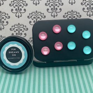 May include: A black tin with a white label that reads "Pin Dreams On Magnetic Pins". The tin contains six magnetic pins, three pink and three light blue.