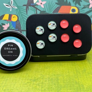 May include: A black tin with a white label that says "Pin Dreams On - Magnetic Pins". The tin contains six small round magnets with a toucan design and six small round magnets in a bright pink color.