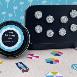 May include: A black metal container holding eight silver glitter magnetic pins. A round tin with the text "PIN DREAMS ON - MAGNETIC PINS" is next to the container. The background has a beach-themed pattern.