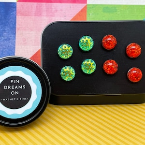 May include: A black tin with a label that reads "Pin Dreams On - Magnetic Pins". The tin contains a set of small, round, magnetic pins with green and red glitter.