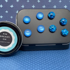 May include: A tin with a blue and white label that says "Pin Dreams On - Magnetic Pins." The tin contains six magnetic pins, three are solid blue and three are blue with glitter.