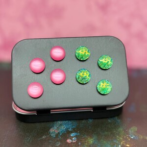 May include: A black rectangular tin with a hinged lid, holding six round magnets. Four magnets are a solid pink, and two are green with yellow and blue accents. The tin is on a pink background.