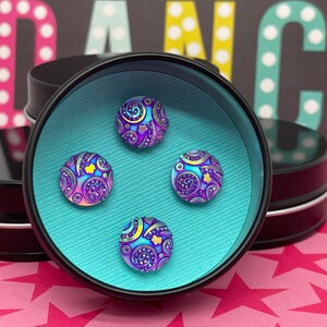 May include: Four iridescent purple and blue buttons with a swirling pattern and gold accents are displayed in a black tin with a turquoise blue interior.