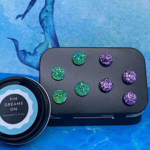 May include: A set of eight magnetic pins in a black tin. The pins are round and have a glittery finish. There are four green pins and four purple pins. The tin has the words "Pin Dreams On" and "Magnetic Pins" printed on it.