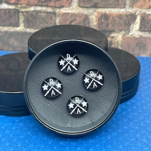 May include: Four black round buttons with a white design of crossed swords and stars. The buttons are arranged in a circle inside a black container.