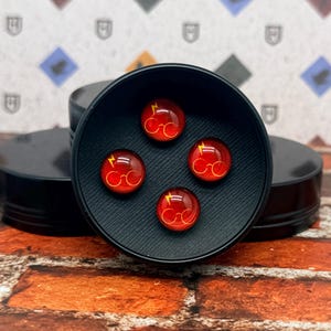 May include: Four red and gold glass cabochons with a lightning bolt and glasses design, displayed in a black container. The cabochons are round and have a glossy finish. The container sits on a brick surface.