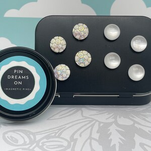 May include: A black tin with a white lid containing a set of magnetic pins. The tin has the words "Pin Dreams On - Magnetic Pins" printed on the lid. The pins are round and have a white iridescent finish. Some of the pins have a textured surface.