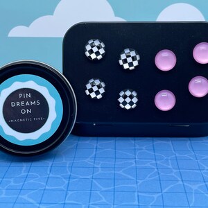 May include: A black tin with a white label that says "Pin Dreams On - Magnetic Pins". The tin contains eight magnetic pins, four are black and white checkered and four are pink.