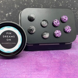 May include: A black tin with a white label that reads "Pin Dreams On - Magnetic Pins". The tin contains eight small, round, magnetic pins. Four pins are black with silver glitter and four pins are purple with silver glitter.
