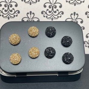 May include: A black tin box with a variety of round, sparkly buttons. The buttons are in two colors: gold and black. The buttons are arranged in two rows of three.