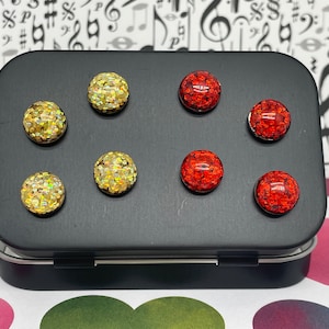 May include: A tin box with eight round, glittery, adhesive backed magnets. There are three gold magnets, three red magnets, and two yellow magnets.