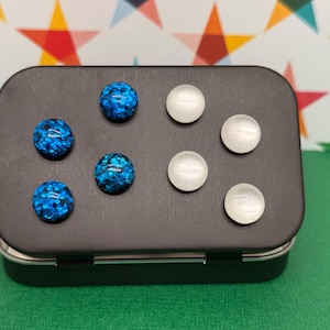 May include: A black rectangular tin with a hinged lid, containing six decorative magnets. Three magnets are blue with glitter, and three are white and opaque. The tin sits on a green surface with a colorful star-patterned background.