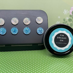 May include: A tin of magnetic pins with a blue and white glitter design. The tin is labeled "Pin Dreams On - Magnetic Pins".