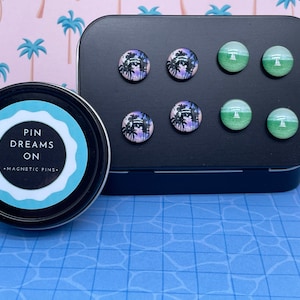 May include: A set of magnetic pins with tropical designs. The pins feature images of palm trees and beach scenes in shades of purple, green, and white. The pins are displayed on a black metal container, and a tin with the text "PIN DREAMS ON - MAGNETIC PINS" is also visible.