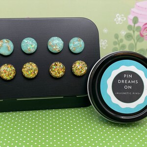 May include: A set of six magnetic pins in a round tin. The pins are decorated with turquoise and gold glitter. The tin has a label that reads "Pin Dreams On - Magnetic Pins".