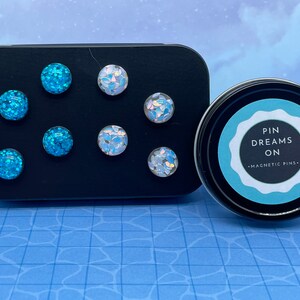 May include: A set of magnetic pins in a black rectangular container and a round tin. The pins are round with a glittery blue or iridescent finish. The tin has the text "PIN DREAMS ON - MAGNETIC PINS" on the label.