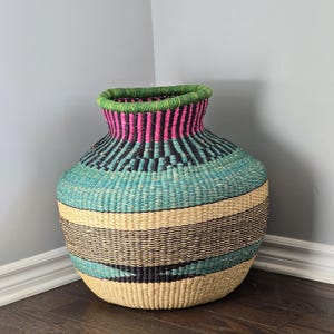 May include: A large, woven basket with a wide base and a narrow neck. The basket features horizontal stripes in natural beige, black, and turquoise, with a pink and green rim. The basket is made of natural fibers.