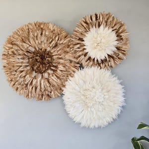 Peut inclure: Trois décorations murales circulaires en plumes. Deux sont de couleur marron clair, et une est blanche. La texture duveteuse crée un aspect doux et naturel. Elles sont fixées sur un mur gris clair, ajoutant une touche de texture.
