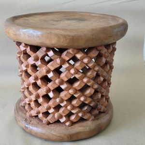 Könnte beinhalten: Handgefertigter Holzschemel mit runder Ober- und Unterseite. Der Korpus weist ein Gittermuster mit geschnitzten geometrischen Formen auf. Der Hocker ist aus hellbraunem Holz gefertigt und hat eine natürliche Oberfläche, die die Holzmaserung zeigt.