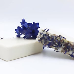 May include: Two bars of handmade soap, one with a white base and the other with a white base and blue and brown flower petals embedded in the soap.