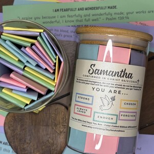 Op de afbeelding: Een glazen pot met een houten deksel gevuld met kleurrijke affirmatiekaarten. De pot is gelabeld met "Samantha" en bevat affirmaties zoals "Sterk" en "Uitverkoren". Een houten hart en extra affirmatiekaarten zijn ook zichtbaar.