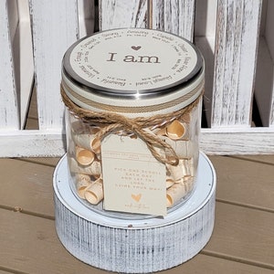 May include: A glass jar filled with rolled up paper scrolls with inspirational words on them. The jar has a metal lid with the words "I am" and a heart on it. The jar is sitting on a white wooden base. The jar is labeled "To bring you hope, comfort, peace".  The jar has a tag that says "Pick one scroll each day and let the Lord guide your way!"
