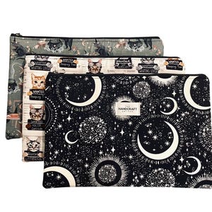 May include: Three zippered pouches with unique patterns. One features a black and white celestial design with moons and stars. Another has cat illustrations with the text "Wanted". The third has a green background with black cat silhouettes.