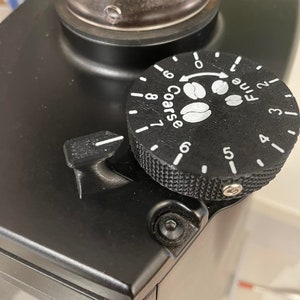 May include: Close-up of a black coffee grinder with a dial that reads "Coarse" and "Fine" with numbers 1-9. The dial is set to 7.